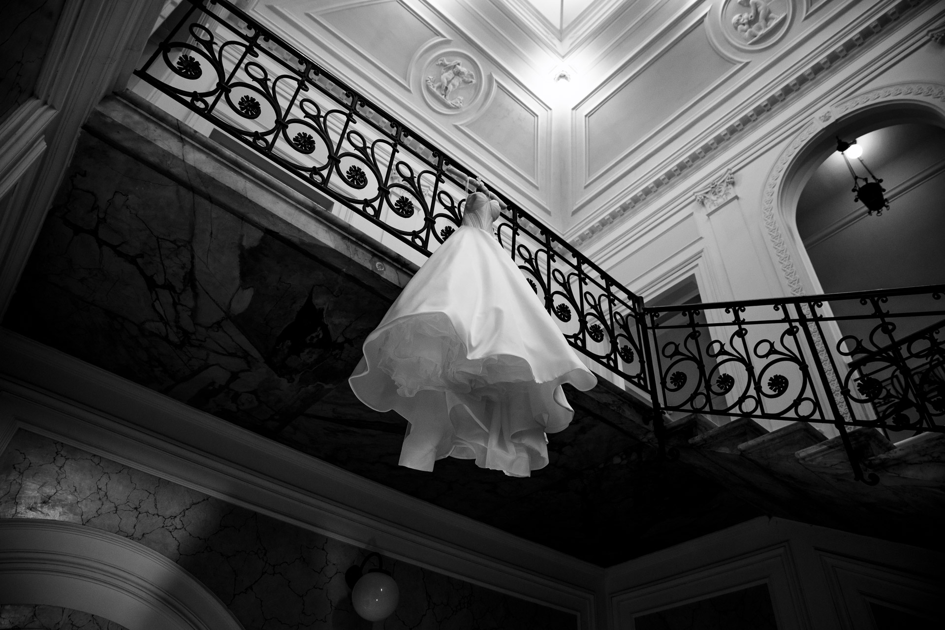 Black-and-white photo of a wedding gown suspended from an ornate iron stair railing in a grand, neoclassical interior.