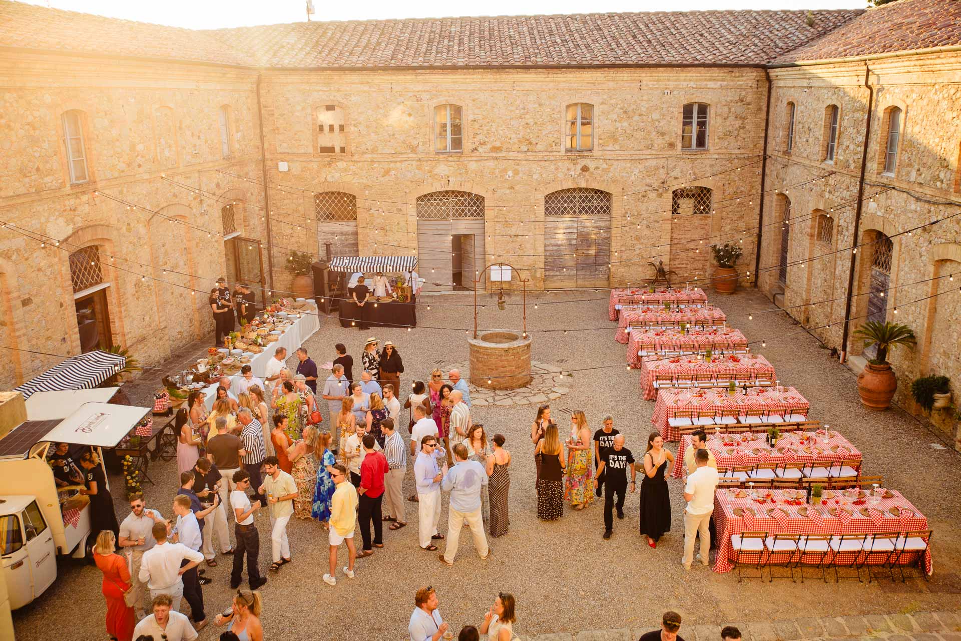 Outdoor courtyard party in a stone building with string lights and long red-and-white checkered tables.