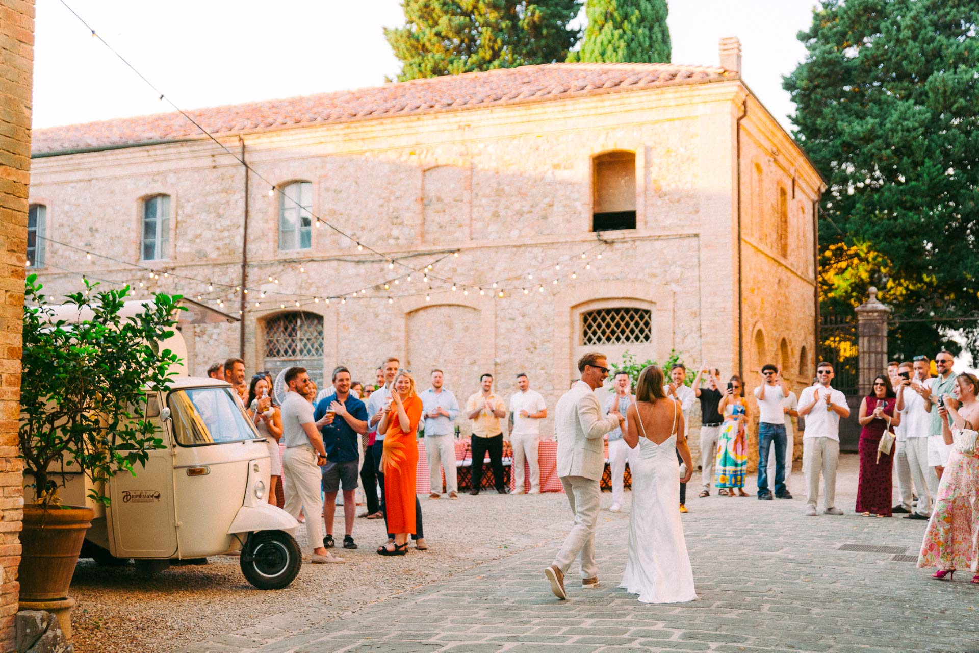 Bride and groom walk toward the camera at an outdoor wedding, guests applauding under string lights in a stone courtyard.
