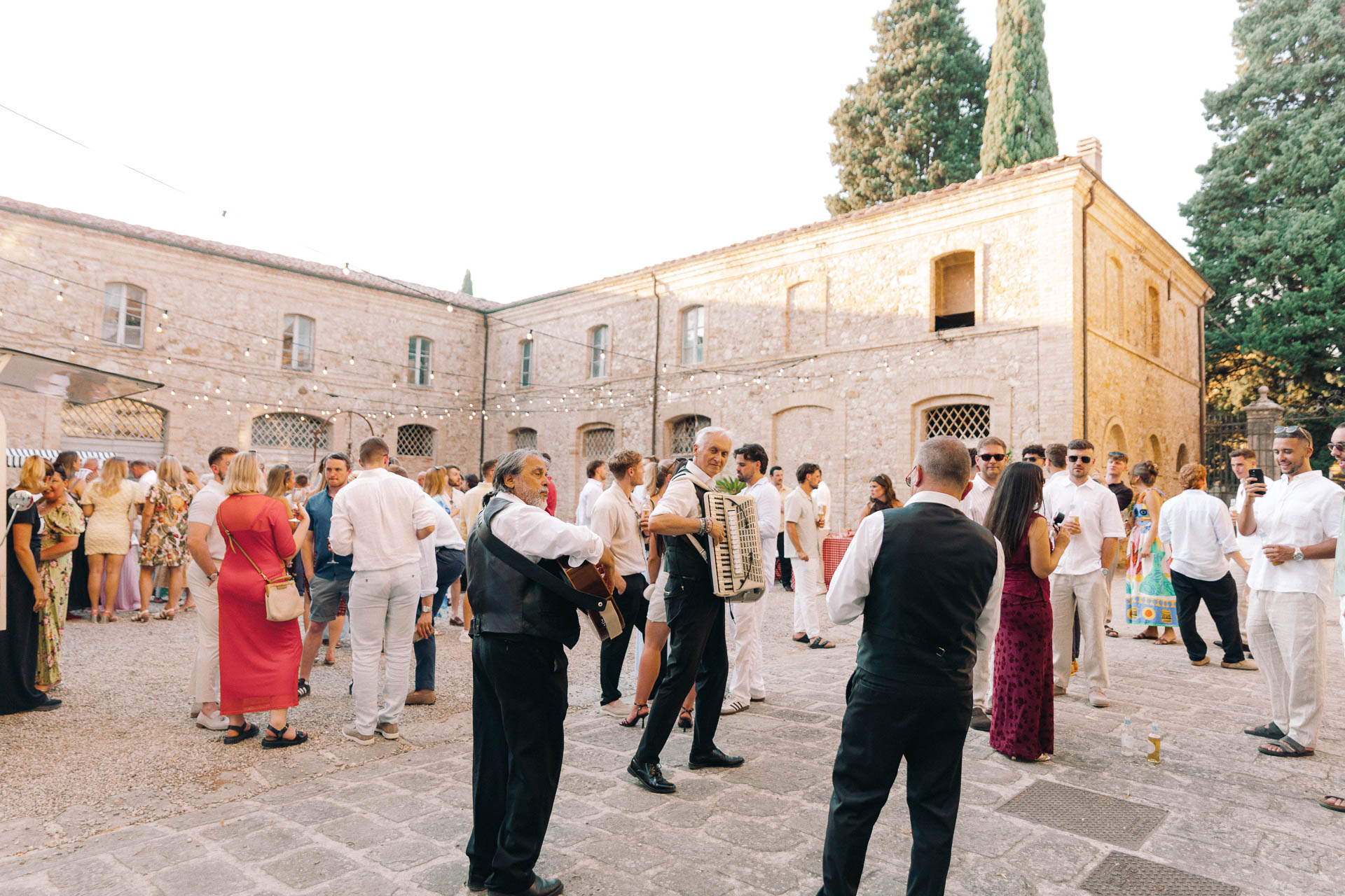 Outdoor courtyard party with guests mingling as two accordions players perform among string lights and a stone building in the background.