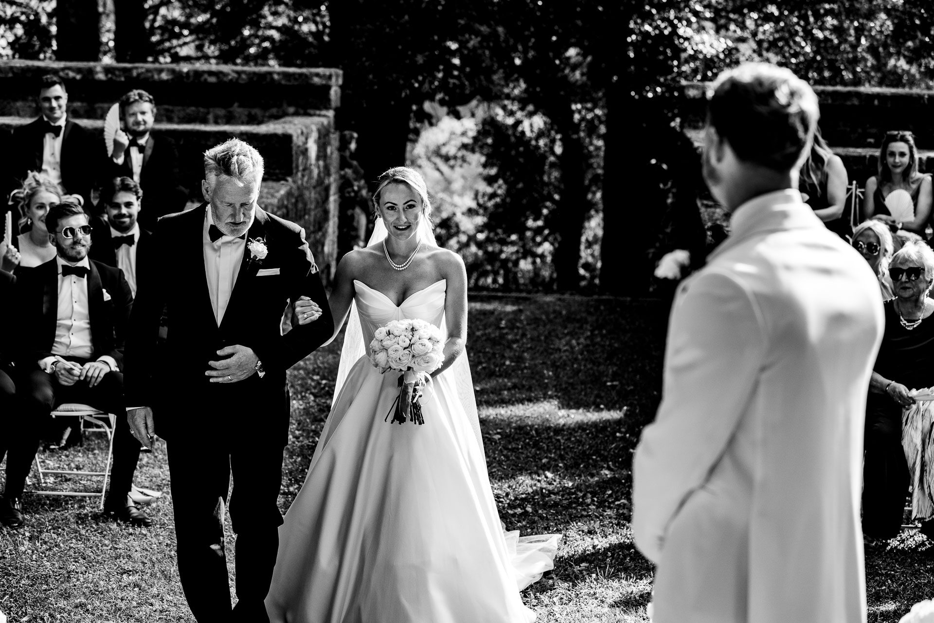 Bride in a white wedding gown walks arm-in-arm with an older man in a tuxedo; bouquet in hand, as guests look on outdoors in the background.