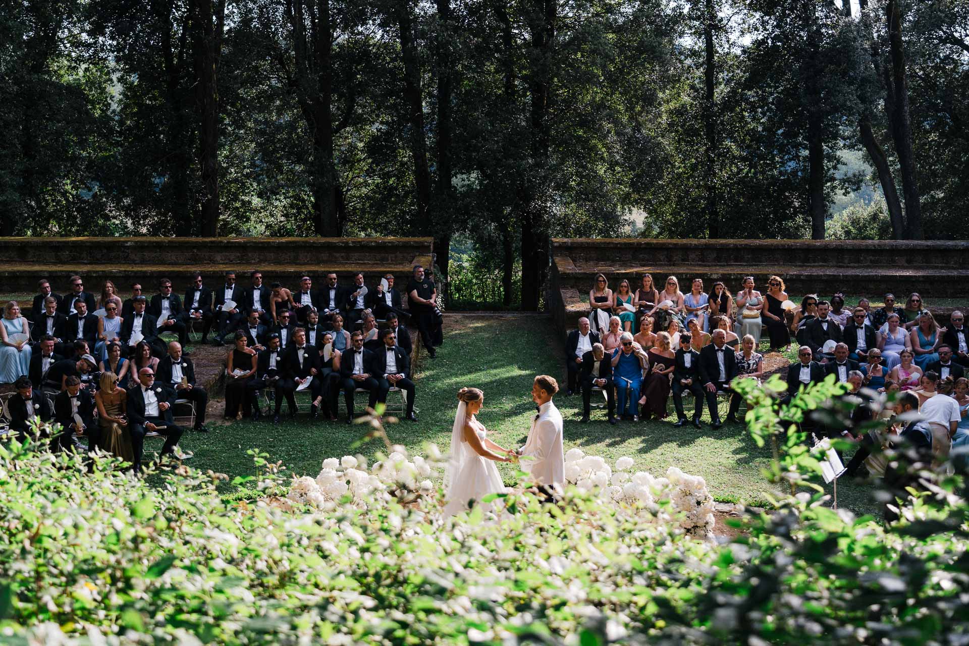 Bride and groom exchange rings on a grassy aisle as guests in formal attire watch in an outdoor stone amphitheater setting.