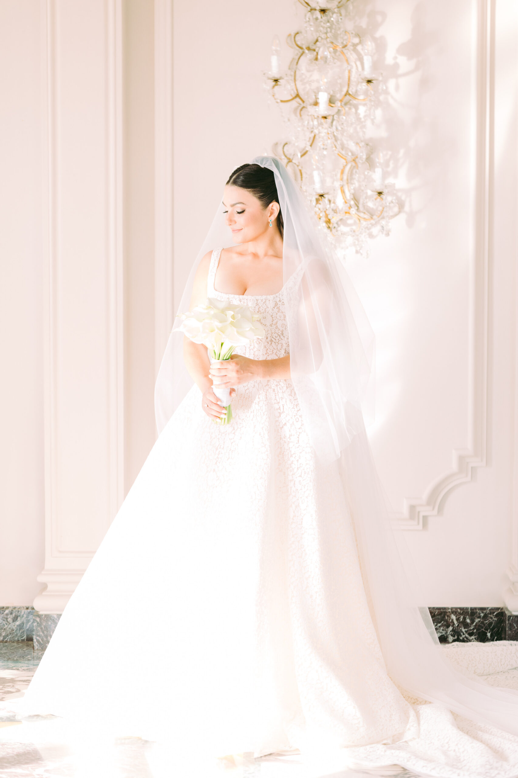 Bride in lace wedding gown with veil, holding a white bouquet of calla lilies, indoors by a crystal chandelier.