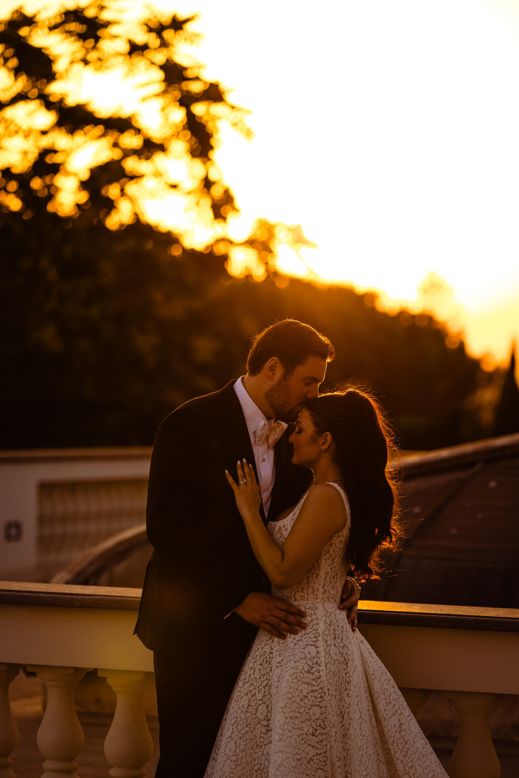Bride and groom embrace on a sunlit balcony as the warm sunset glows behind them, creating a romantic silhouette.