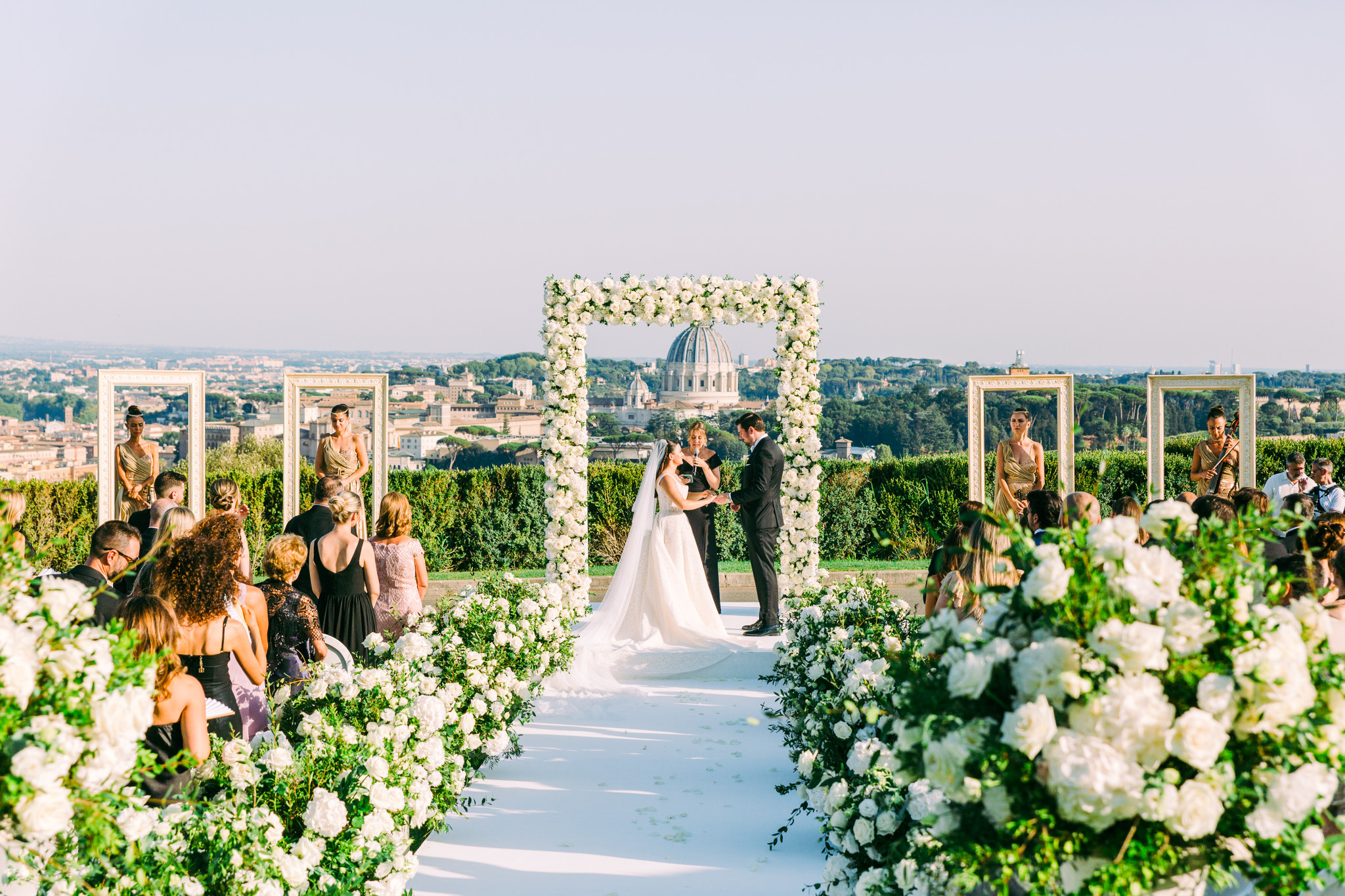 Bride and groom exchange vows beneath a white floral arch at an outdoor rooftop ceremony, with guests seated along a flower-lined aisle.