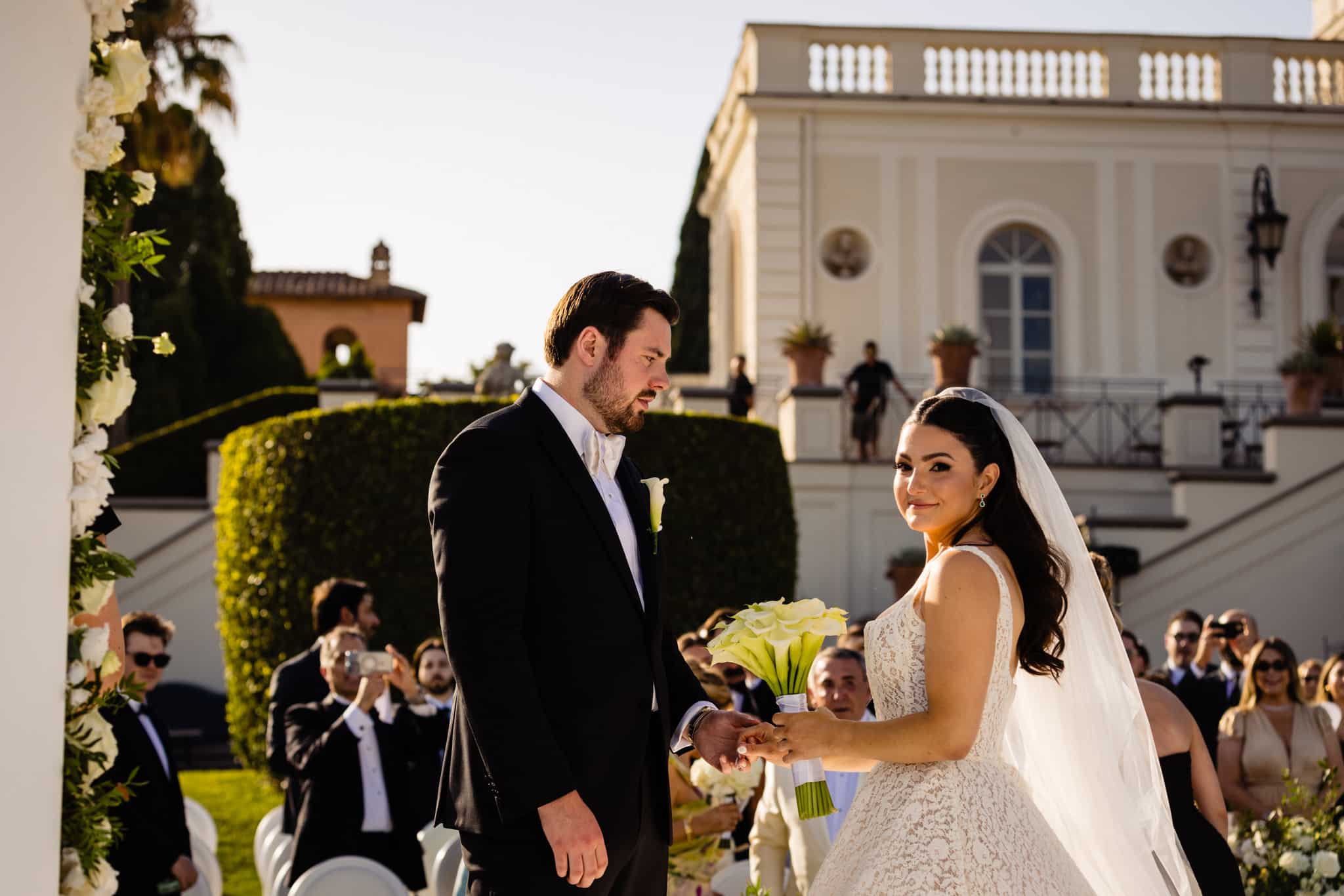 Bride in a lace wedding dress and groom in a dark tux exchanging rings outdoors, with guests and a cream villa in the background.
