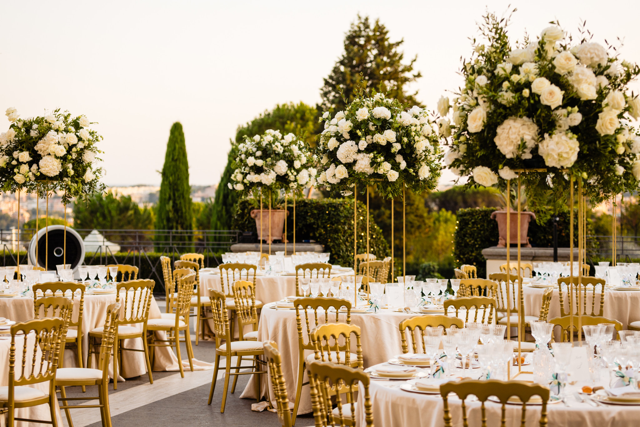 Outdoor wedding reception with tall white floral arrangements on gold stands above round tables set with beige linens and gold chairs beneath a warm sky.